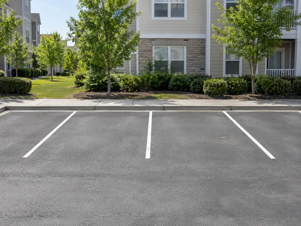 A realistic photo of four adjacent empty parking spaces in a small neighbourhood parking lot, with residential buildings visible farther along a level sidewalk. No signs, text, logos or identifiable real places.