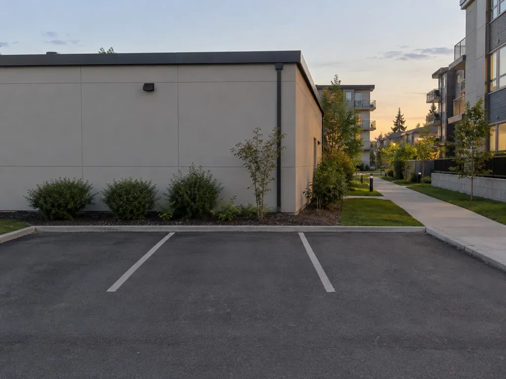 A realistic photo of three empty parking spaces behind a small office building at dusk, with a paved pedestrian route continuing toward nearby condos. No signs, text, logos or identifiable real places.