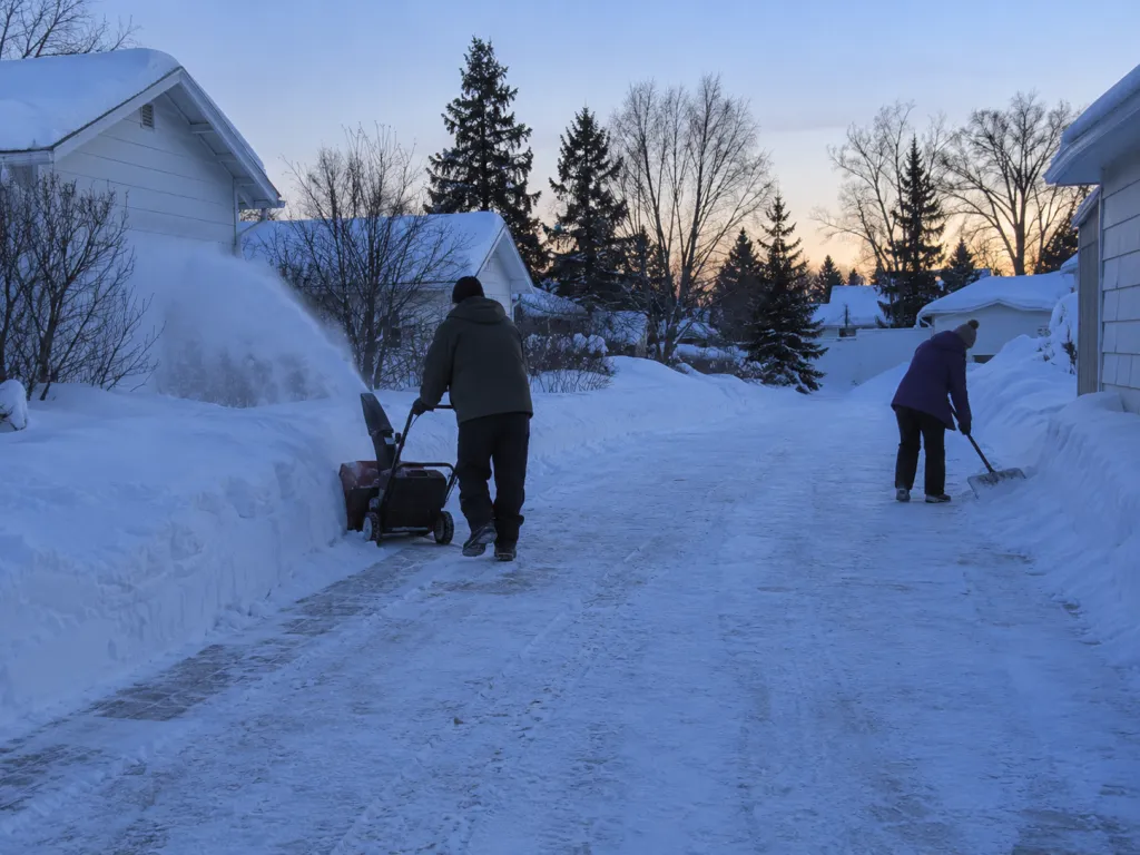 A realistic photo of two adults clearing a private residential lane before dawn, one operating a snowblower while the other clears the edges with a shovel. No text, logos or identifiable real places.